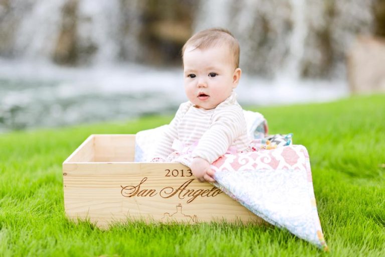 cutest little baby girl in middle of grass waterfall behind sitting clueless in wooden wine box with blanket during infant photoshoot with danielle jacqueline in byhalia mississippi