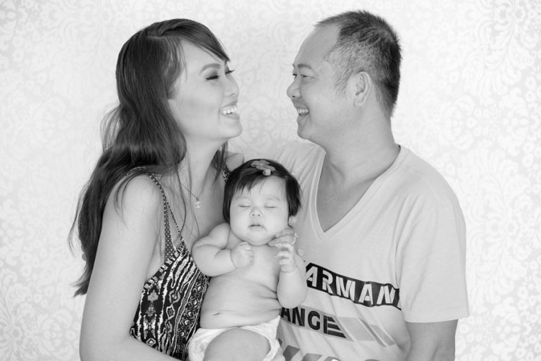 mom and dad looking at each other laughing black and white baby portraits with newborn daughter in middle seated in their arms custom backdrop photographer studio danielle jacqueline mississippi