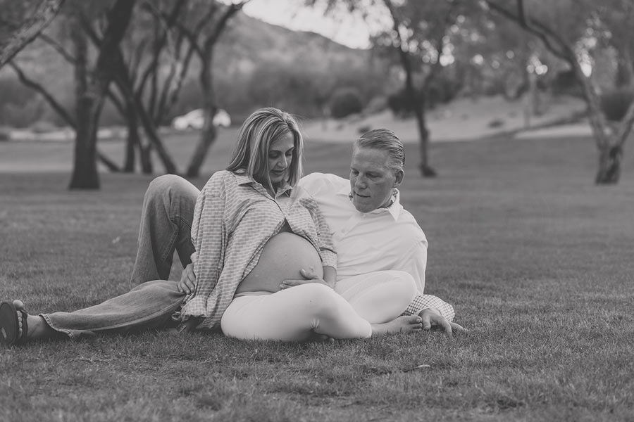 wife looking down at pregnant belly exposed by button up shirt seated in grass with spouse laying behind her imagining what parenting will be like as they await their new arrival photo by danielle jacqueline in mississippi desoto county