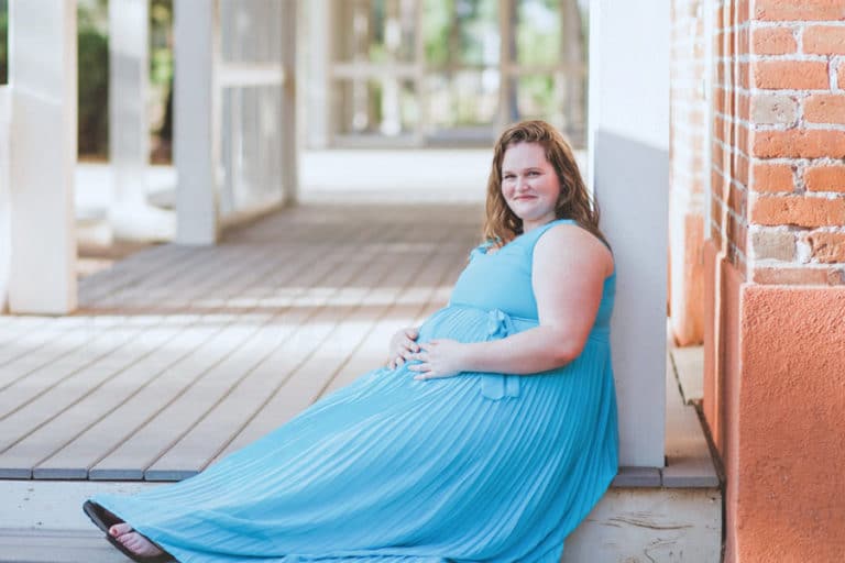 red haired woman expecting child seated on plantation porch in north mississippi farmhouse maternity portraits bright blue long dress over wooden steps curls matching red brick residence while leaning on white pillars outside in desoto county ms