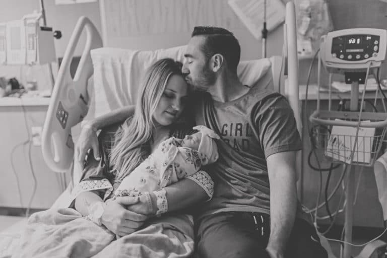 kissing wife on forehead good job delivering healthy baby girl on reclining hospital bed with medical equipment around for black and white photo of new member of family by photographer in olive branch ms