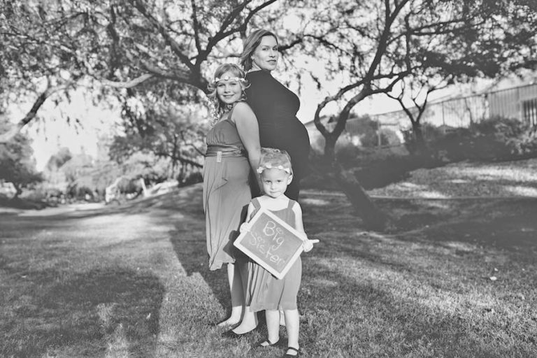 single mother family standing outside residence under tree holding "it's a girl" sign written in chalk by youngest daughter in dress stunning black and white maternity portrait by danielle jacqueline photography