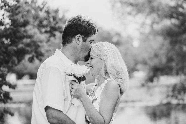 anniversary portrait of happy husband kissing bride's forehead black and white with flowered trees next to pond in love by danielle jacqueline mississippi wedding photographer