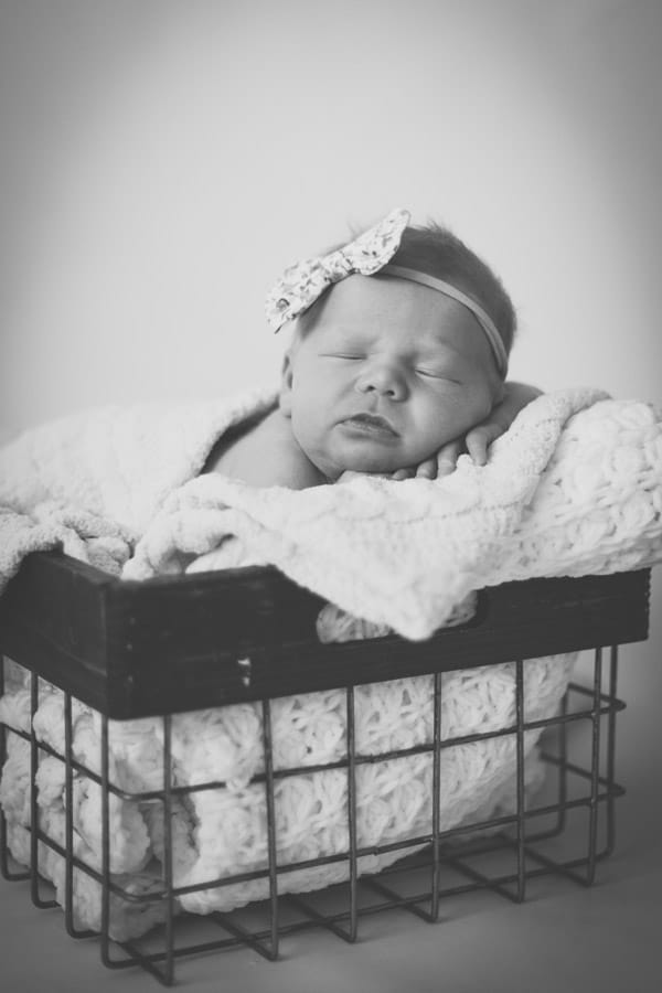 black and white portrait of infant baby girl peeking head over side of metal wire bin with blanket and flower headband sleeping for mom and daddy in olive branch ms by danielle jacqueline photography