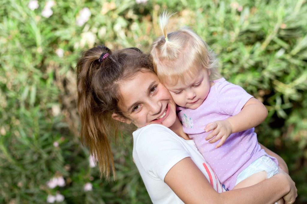 toddler baby girl held by older sibling outside for family photography session at olive branch ms venue with green shrubs in background bright natural lighting big smiles