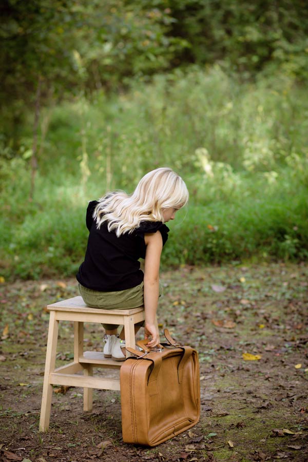 5 year old daughter seated on wooden step stool facing mississippi forrest with old brown leather suitcase waiting to be picked up by her imagination during photoshoot in olive branch with danielle jacqueline at 2 acre venue