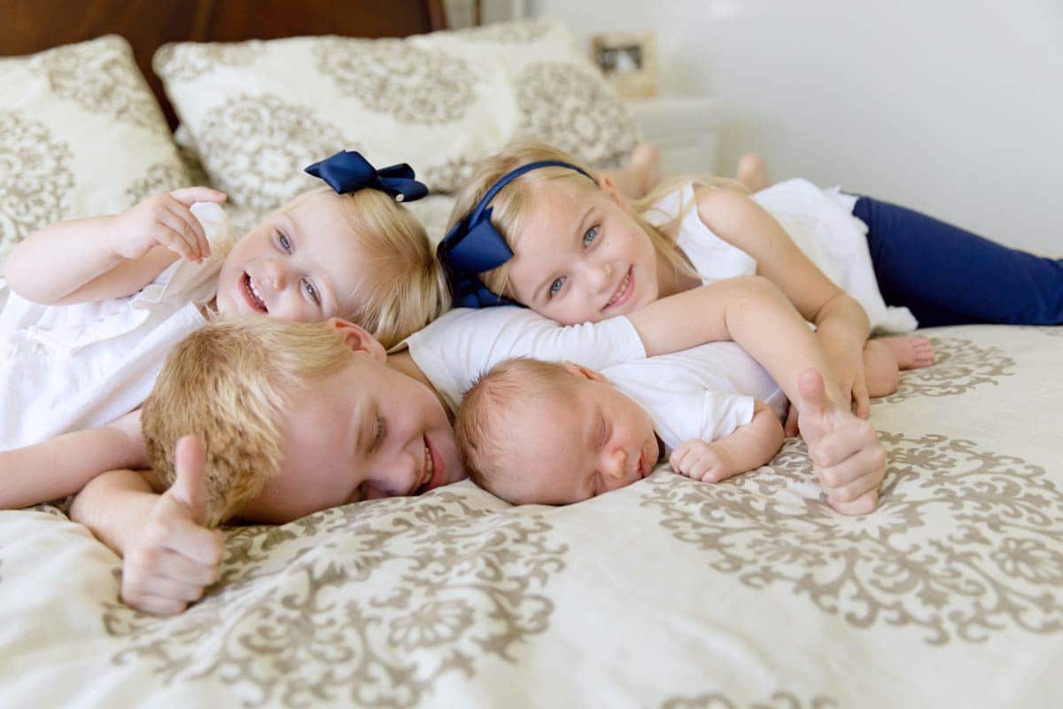 professional portrait of 4 siblings under 8 rolling on bed with newborn baby brother for lifestyle photo session with danielle jacqueline in memphis tennessee