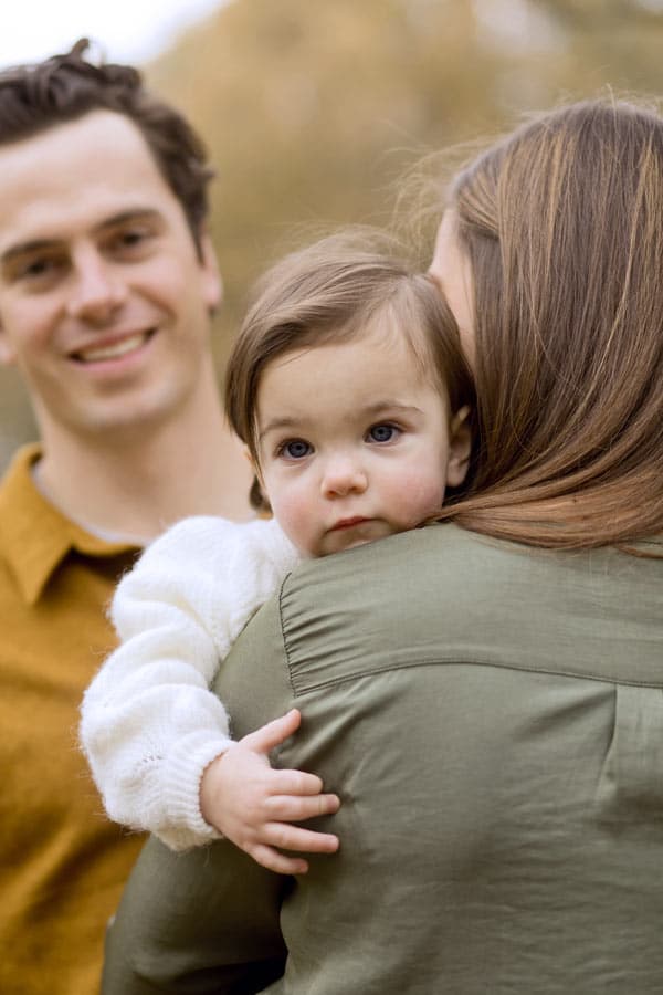 adorable 1 year old clinging to mother's arm with daddy looking from behind in beautiful autumn setting in olive branch ms with danielle jacqueline photography for extended families