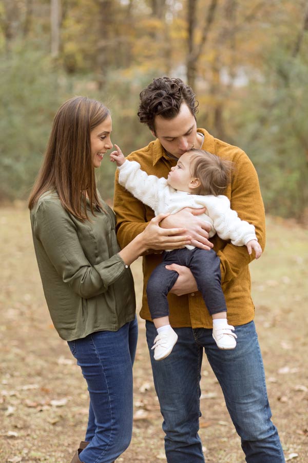 little daughter reaching for mommies nose as daddy holds her tight and kisses forehead autumn colors by trees at photography venue in olive branch ms