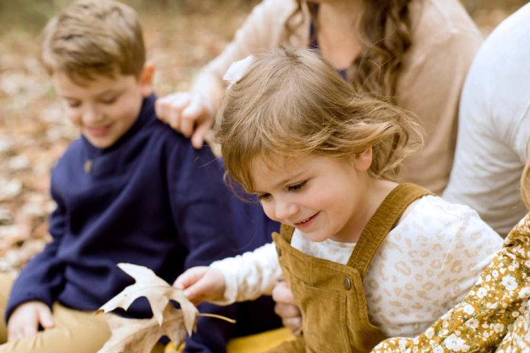 young daughter toddler eager to leave parents lap with autumn leaf yellow older brother in sweater onlooking for extended family photography session in desoto county by danielle jacqueline