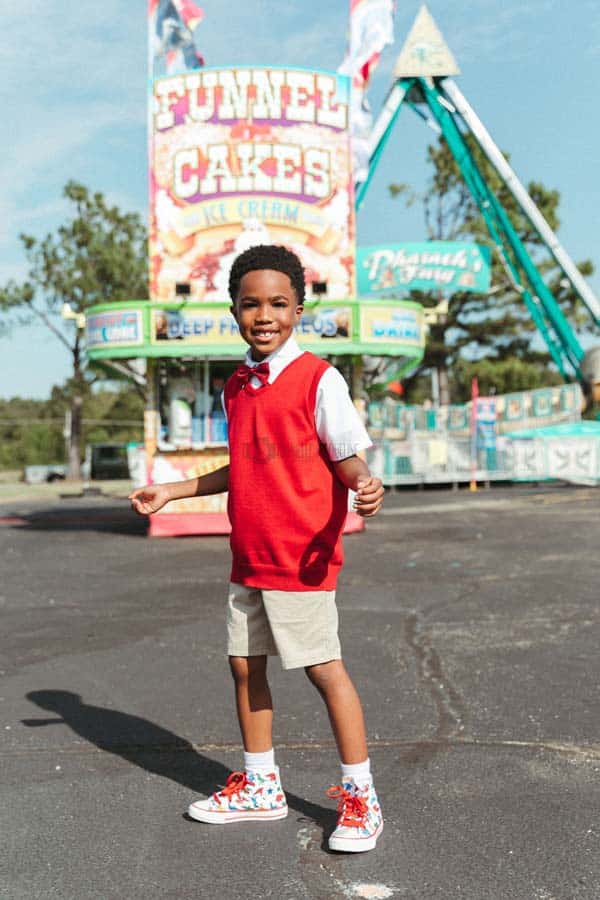 young boy wearing red wool vest and khakis during photoshoot with danielle jacqueline for midsouth fair in southaven ms with rides in background excited family things to do