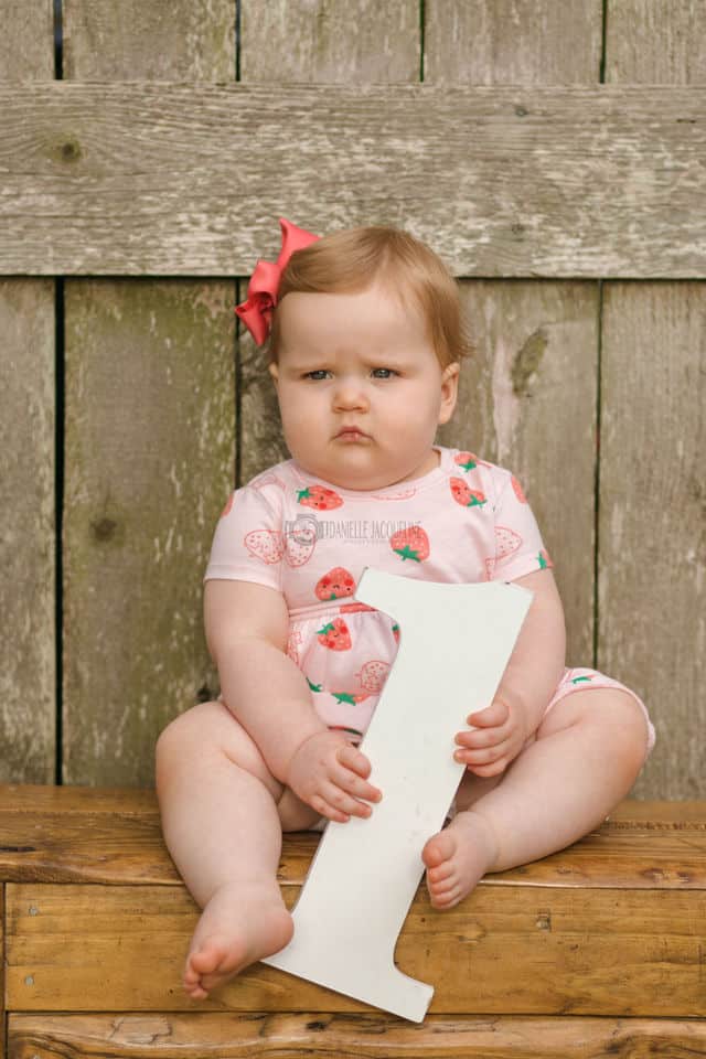 adorable one year old birthday photos baby girl holding one wearing pink flowered onesie seated on wooden stool with wood slat backdrop outdoors with danielle jacqueline kids photographer near memphis tn