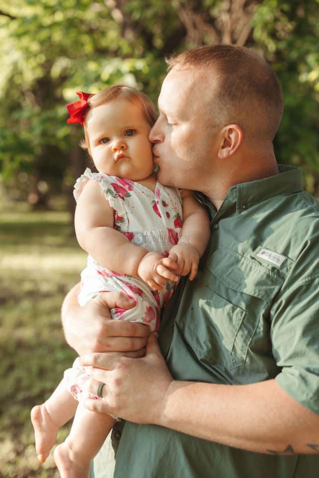 father of one child daughter giving her love on chubby cheeks holding her tightly with warm glow from sun setting in mississippi backyard desoto county photographer