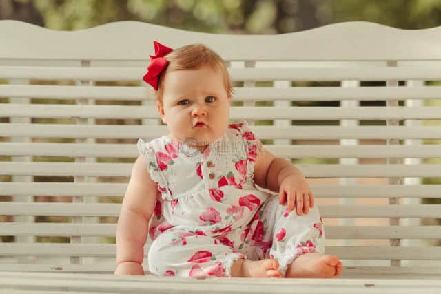 grumpy baby girl turning one warming up to the camera while swinging on a porch swing cute pose pink flowered sun dress with bow professional child photographer olive branch ms