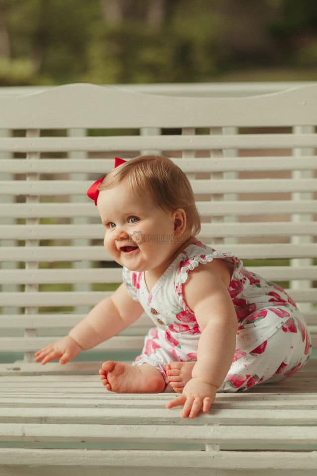 laughing baby girl one year old on porch swing engaged with parents behind photographer danielle jacqueline during parent session in olive branch mississippi