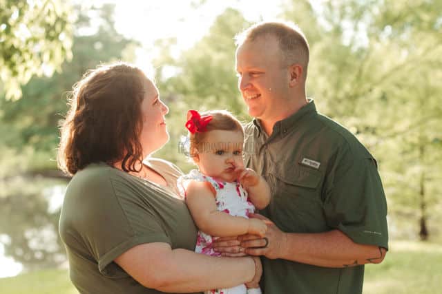 father looking to left into mother's eyes both parents holding baby girl with hand in mouth beautiful sunlit glow behind them in front of natural body of water muggy day first birthday